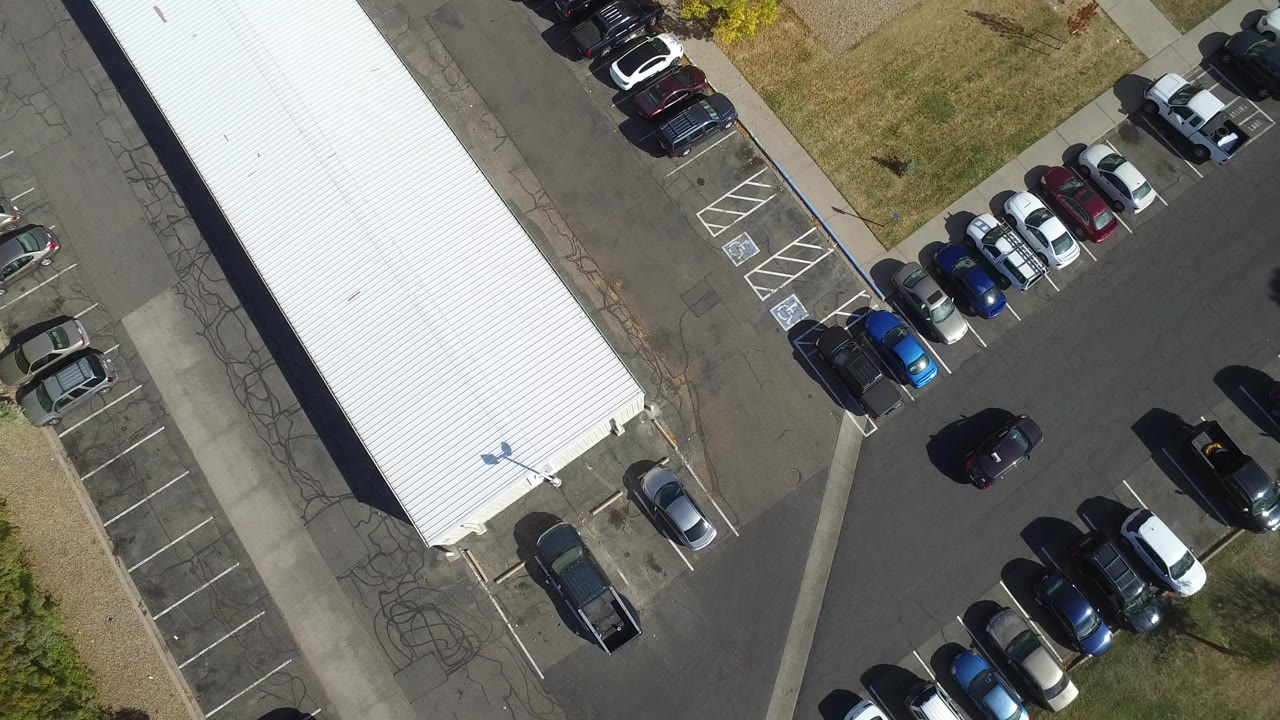Black car crosses the center of an asphalt parking lot from an overhead angle.
