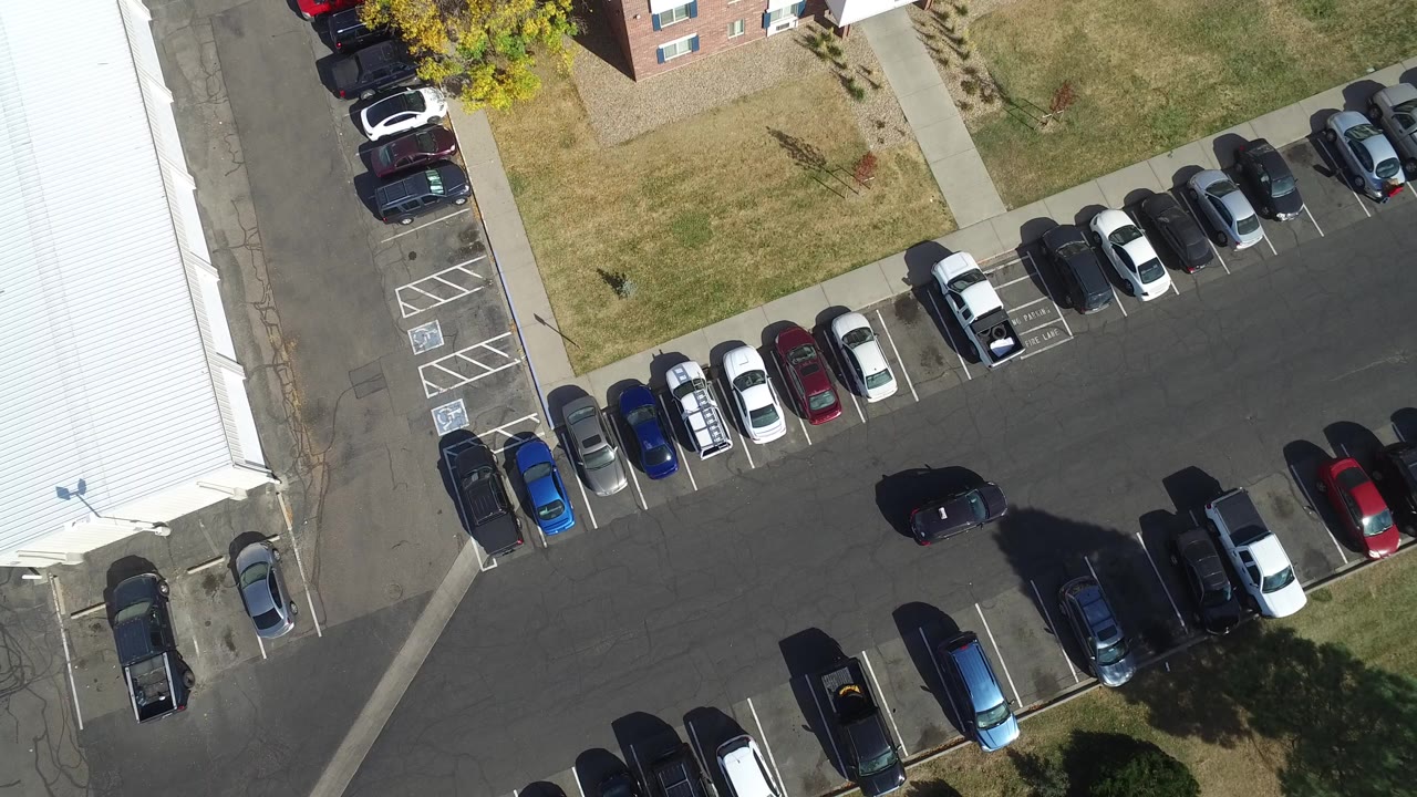 Top-down frame in Hidden Lake, Colorado showing a black car near the middle of a parking lane, rows of parked cars, white-roofed buildings, grass, and no visible people.