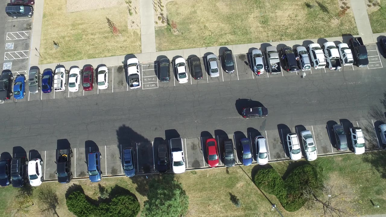 Hidden Lake, Colorado parking lot seen from above, with the dark car slightly farther along the center lane between dense rows of parked cars and sunlit grass borders.