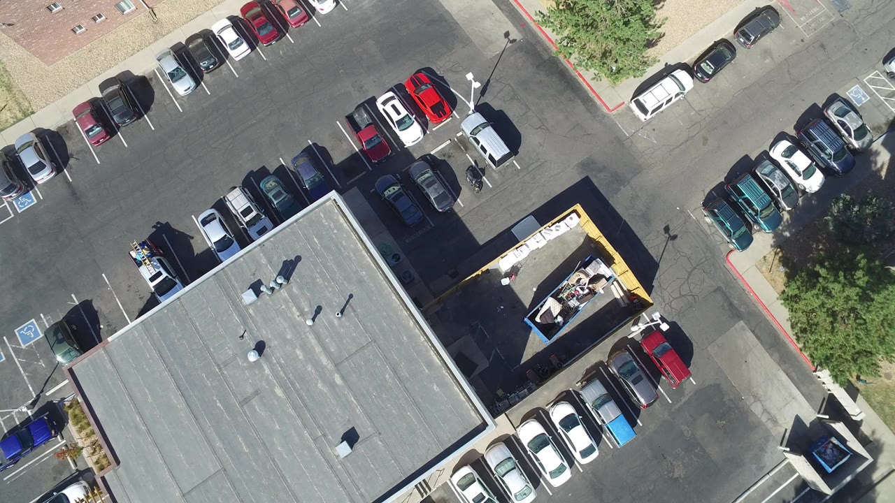Aerial frame over a Hidden Lake, Colorado commercial parking lot with a gray rooftop, parked cars, asphalt lanes, trees, and no visible people.