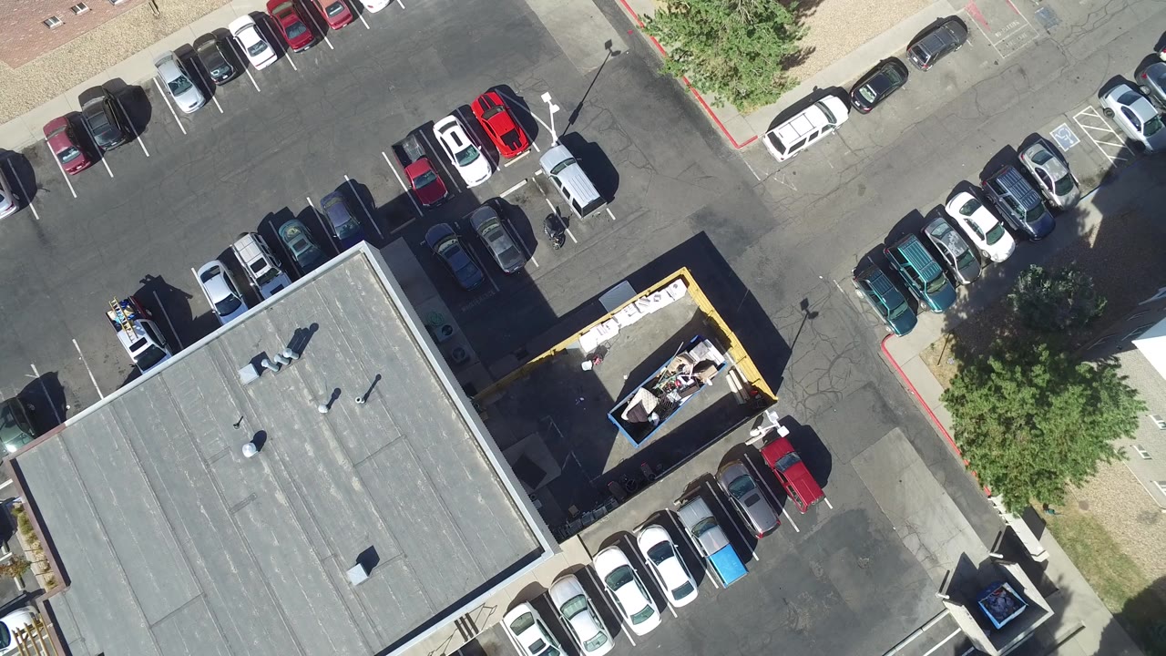 Aerial parking lot frame with a rooftop, parked vehicles, and sidewalk edges.