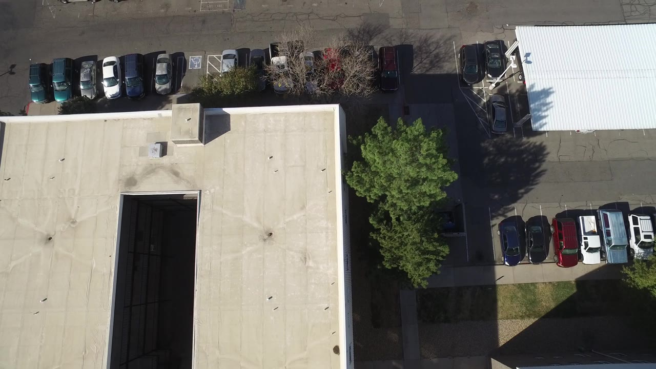 Overhead frame in Hidden Lake, Colorado with the tan building roof centered, rows of parked cars above and at right, a leafy tree, asphalt lanes, and no visible people.