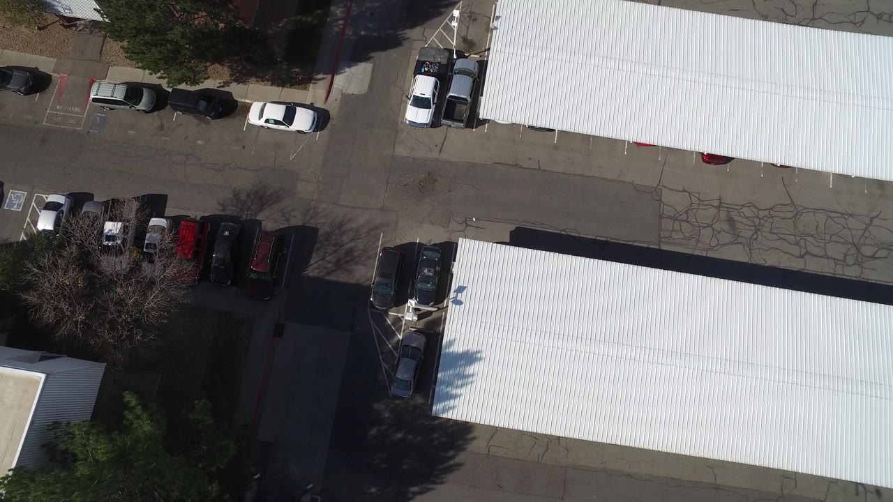 Near-vertical drone frame in Hidden Lake, Colorado with long white roof panels, cracked asphalt, several parked vehicles, tree shadows, and no visible people.