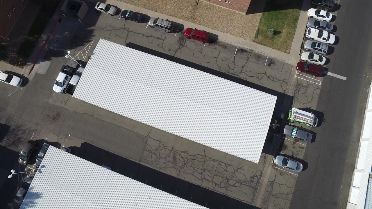 Straight-down aerial frame in Hidden Lake, Colorado showing two white metal rooftops, dark asphalt parking areas, a vertical road, parked cars, and no visible people.