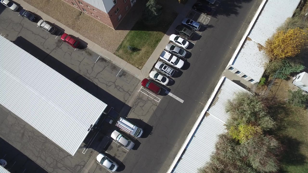 Overhead view of parked cars beside white-roofed buildings and an asphalt street.