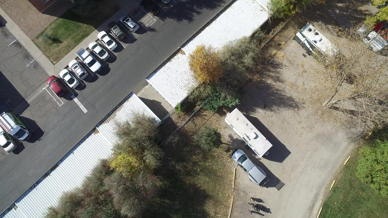 High overhead frame in Hidden Lake, Colorado showing a parking lot with rows of cars at upper left, white-roofed buildings, a white truck or trailer, a silver car, trees, and tiny distant people near the lower edge.