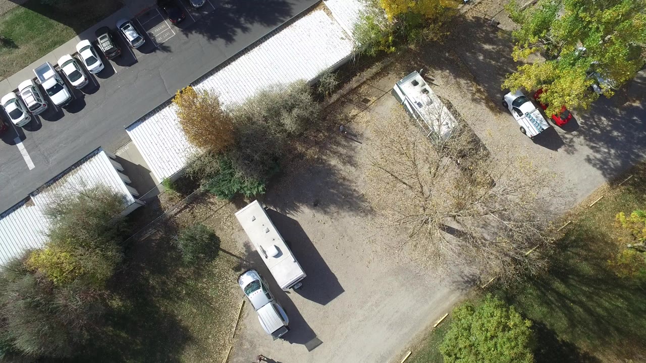 Overhead view of parked cars, storage roofs, an RV, trees, and pavement.