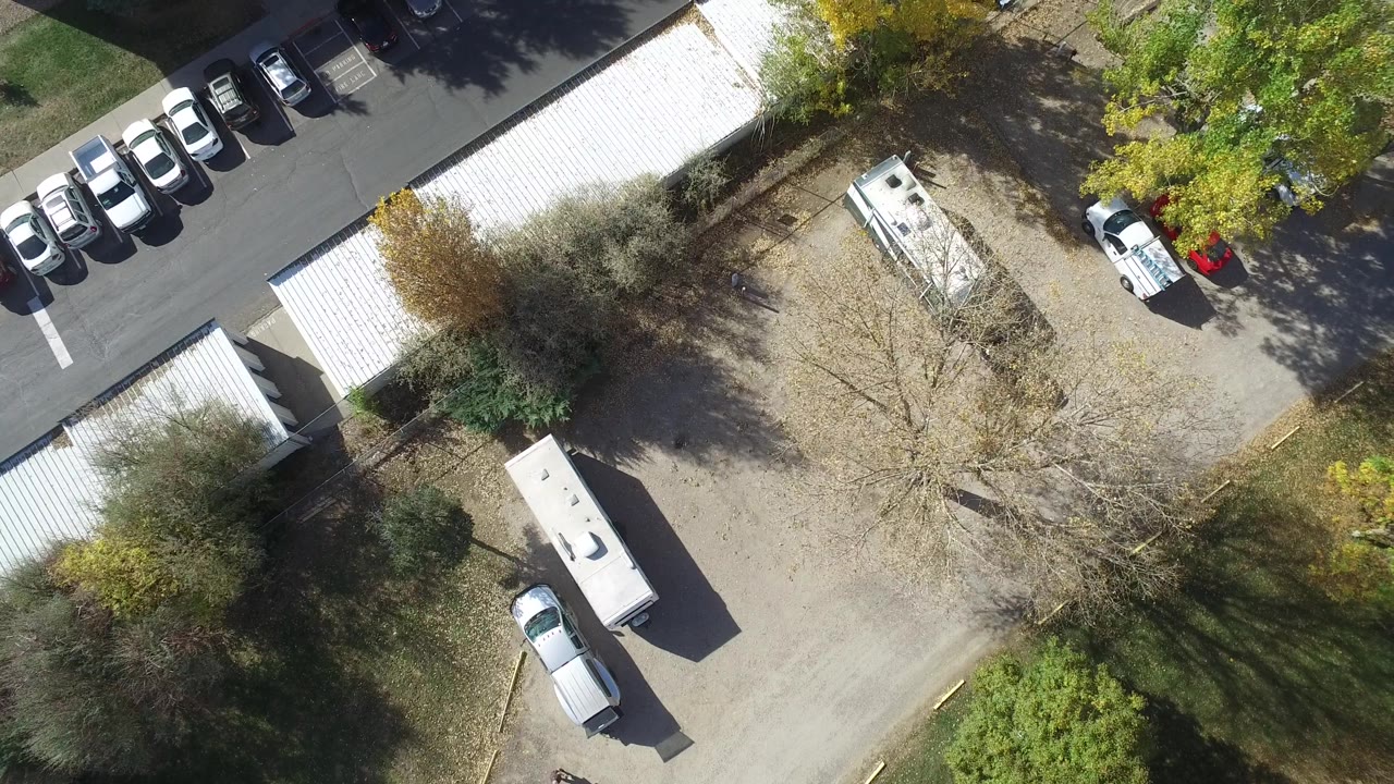 Hidden Lake, Colorado aerial frame with an RV near a gravel lot, parked cars beside a paved lane, white storage roofs, trees, and no visible people.