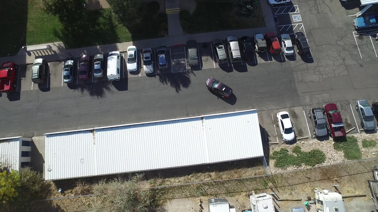 Top-down Hidden Lake, Colorado frame with the white roof centered low, rows of cars along the upper curb, a dark car in the drive lane, and small figures near the bottom edge.
