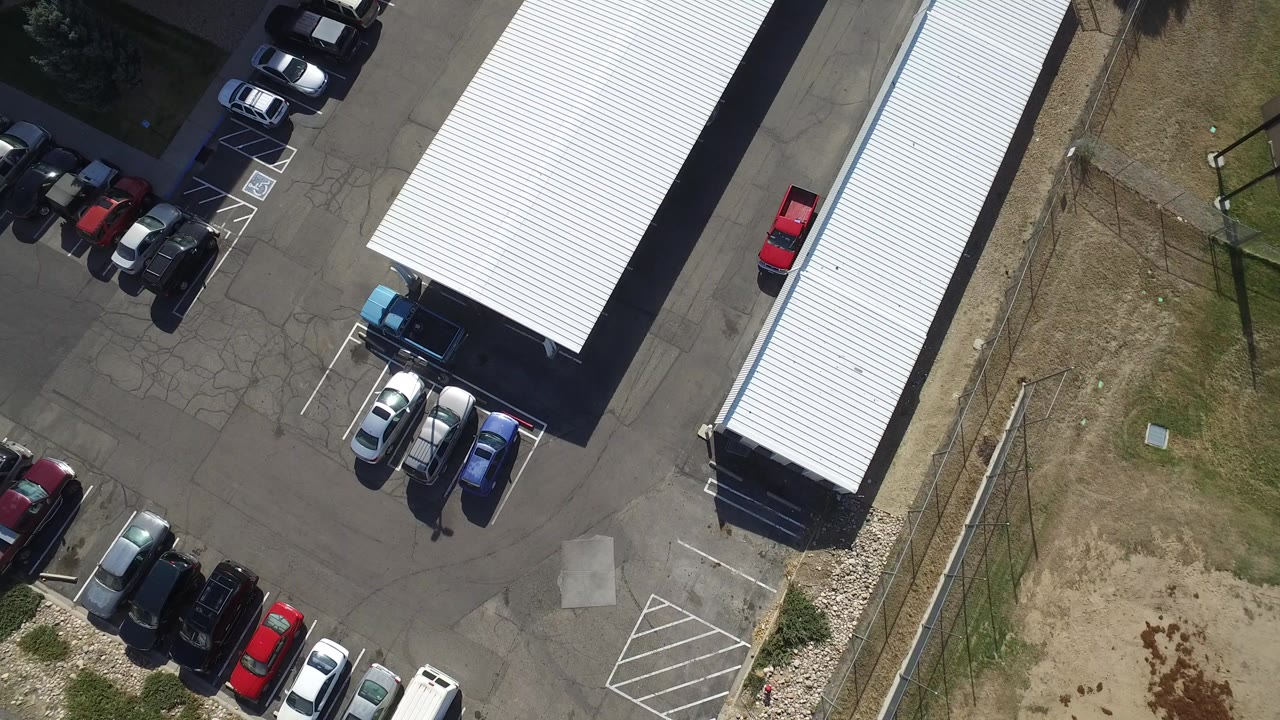 Top-down aerial frame in Hidden Lake, Colorado showing two long white-roofed buildings, parked cars, asphalt drive lanes, dry grass, and no visible people.