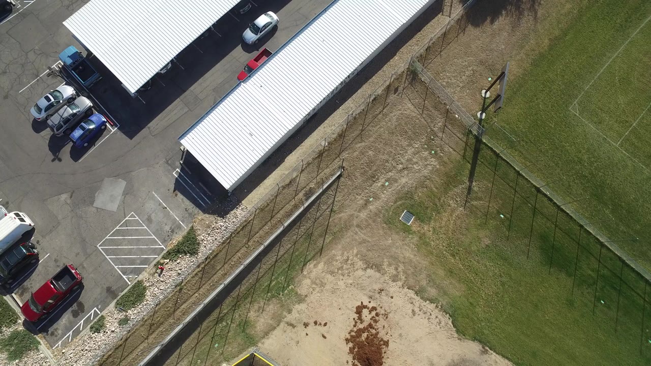 Opening overhead frame in Hidden Lake, Colorado showing a white-roofed warehouse, parked cars, asphalt, fence line, grass, dirt patches, and no visible people.