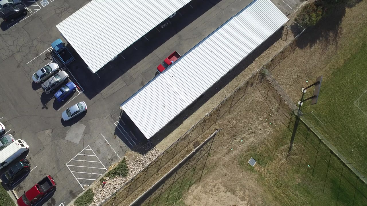 Overhead view of covered parking roofs, parked cars, asphalt, grass, and fencing.