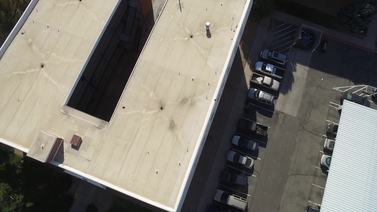 High-angle drone frame over Hidden Lake, Colorado showing pale flat apartment roofs, a shadowed inner gap, parked cars along pavement, and no visible people.