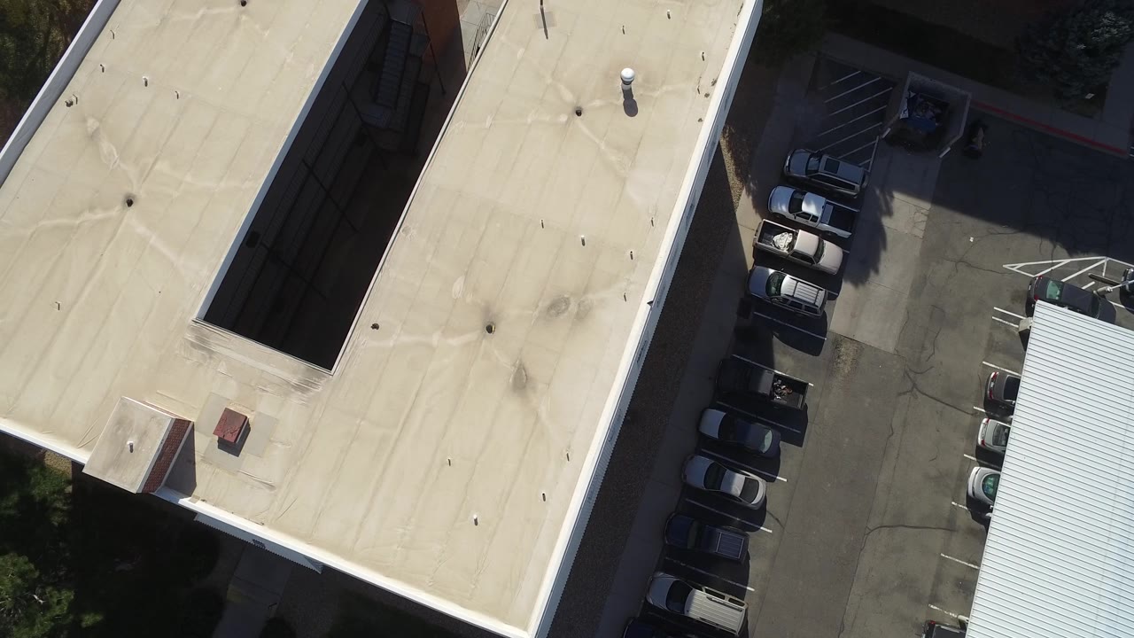 Aerial frame in Hidden Lake, Colorado with a pale roof dominating the left side, parked cars and marked spaces on the right, and no visible people.