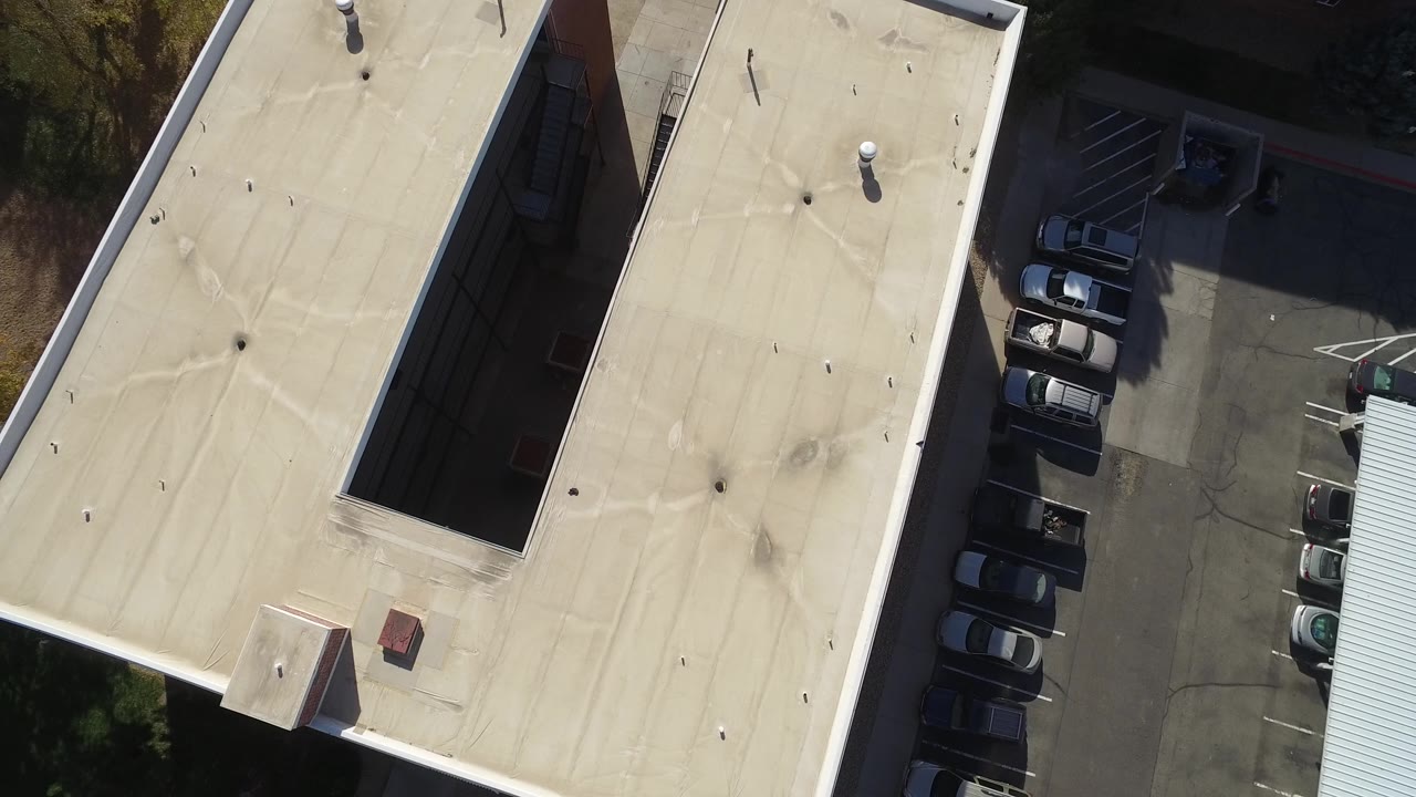 Drone view of apartment roofs, a narrow courtyard gap, and parked cars.