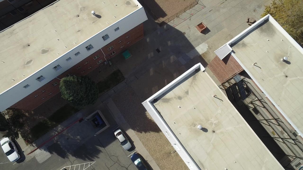 Top-down view of flat-roof buildings, parked cars, trees, and pavement.