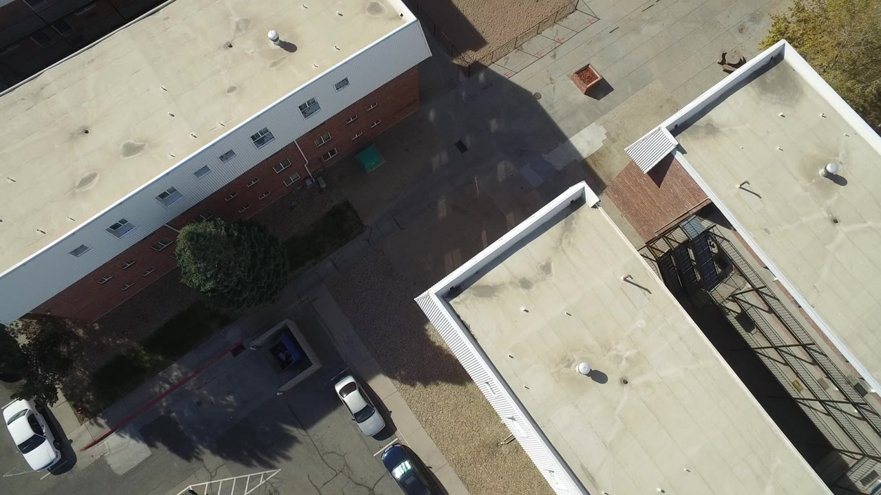 Aerial overhead frame in Hidden Lake, Colorado, showing a long brick building, pale roof surfaces, parked cars along the lane, tree shadows, and no visible people.