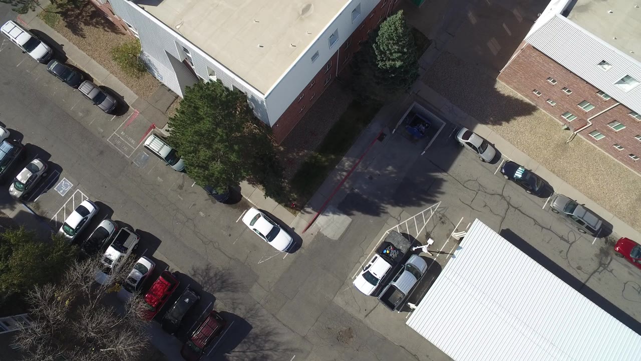 Overhead frame from Hidden Lake, Colorado showing light industrial roofs, parked cars along asphalt lanes, tree shadows, and no visible people.