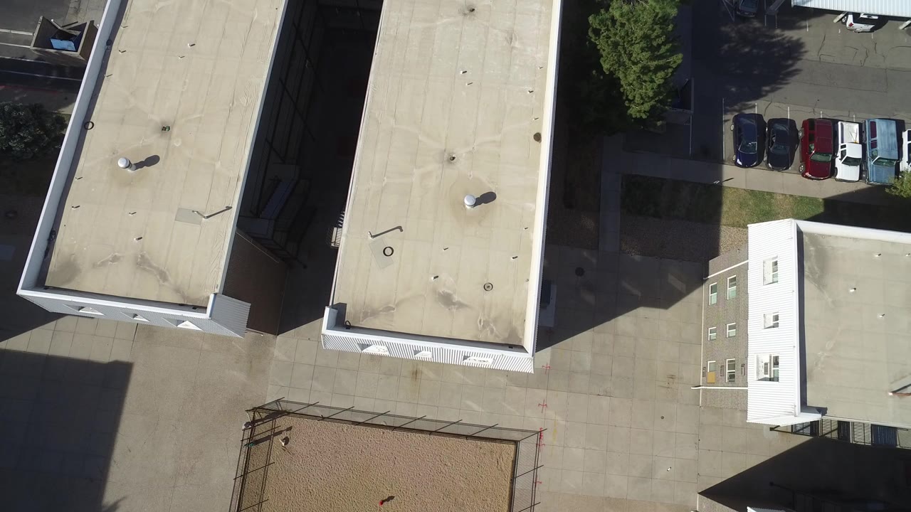 Overhead view of apartment rooftops, paved courtyard, parked cars, and a fenced sand area.