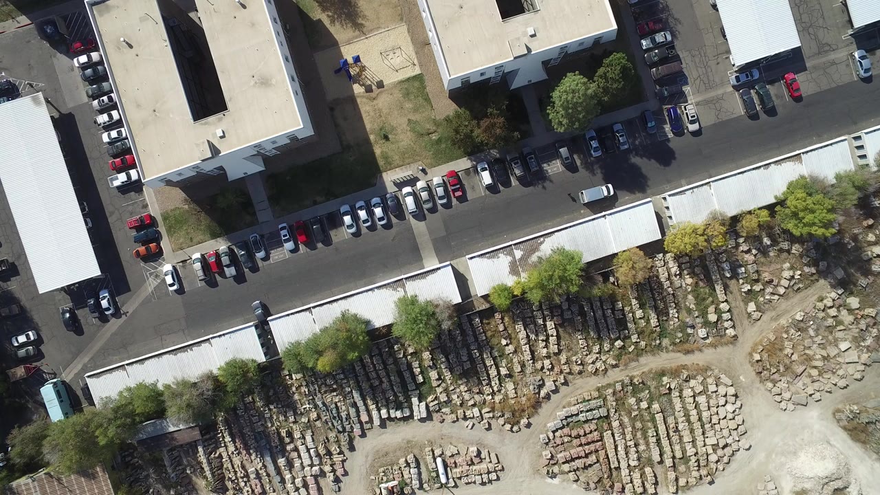 Second overhead frame in Hidden Lake, Colorado with the parking row and white-roof building centered above a storage yard of stacked stone or construction materials, with no visible people.