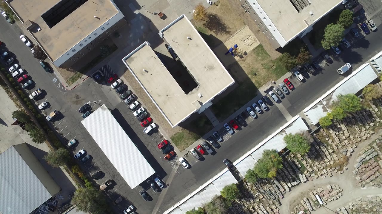 Top-down aerial frame over Hidden Lake, Colorado showing flat-roof buildings, parked cars along two streets, trees, and a materials yard with no visible people.