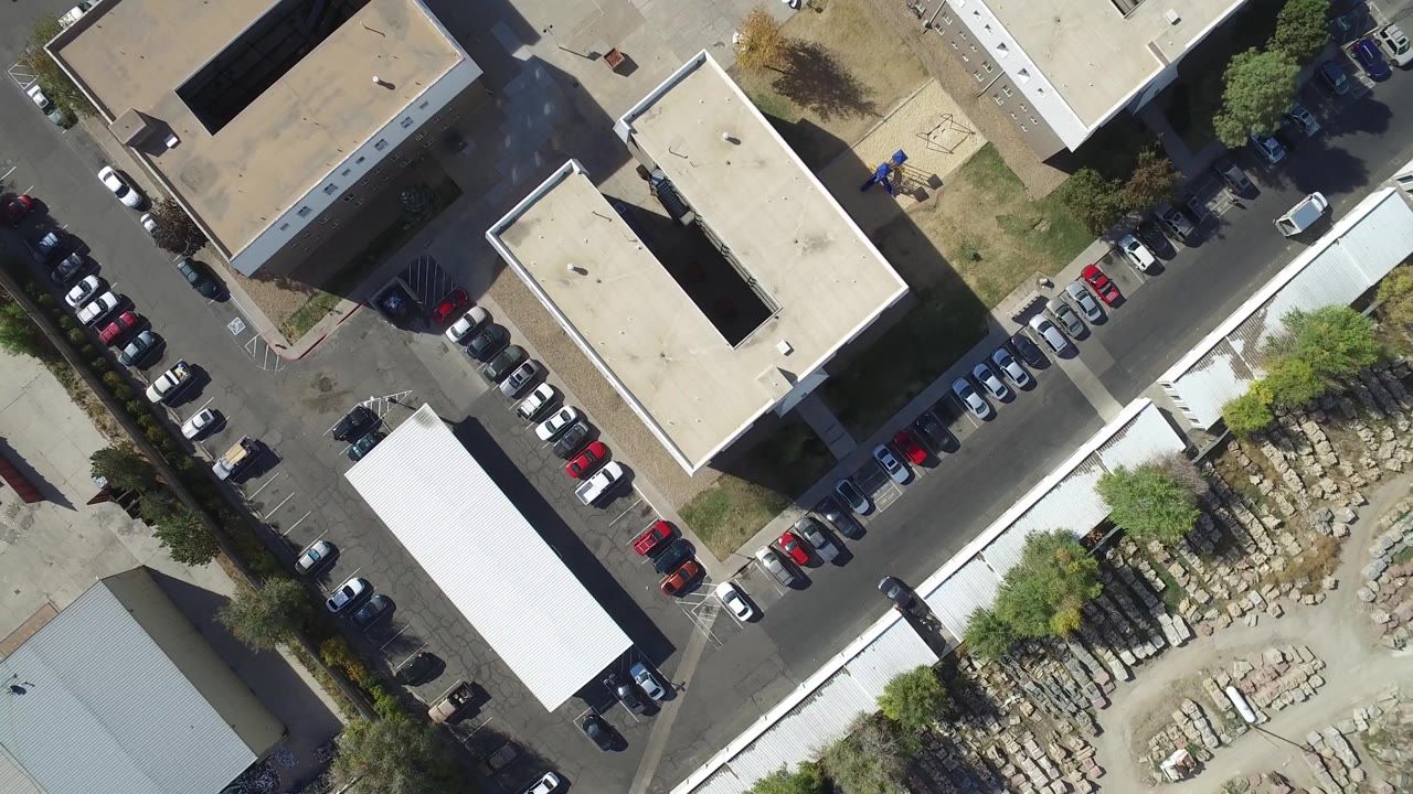Vertical drone view of tan buildings, parked cars, a street corner, trees, and an open yard.