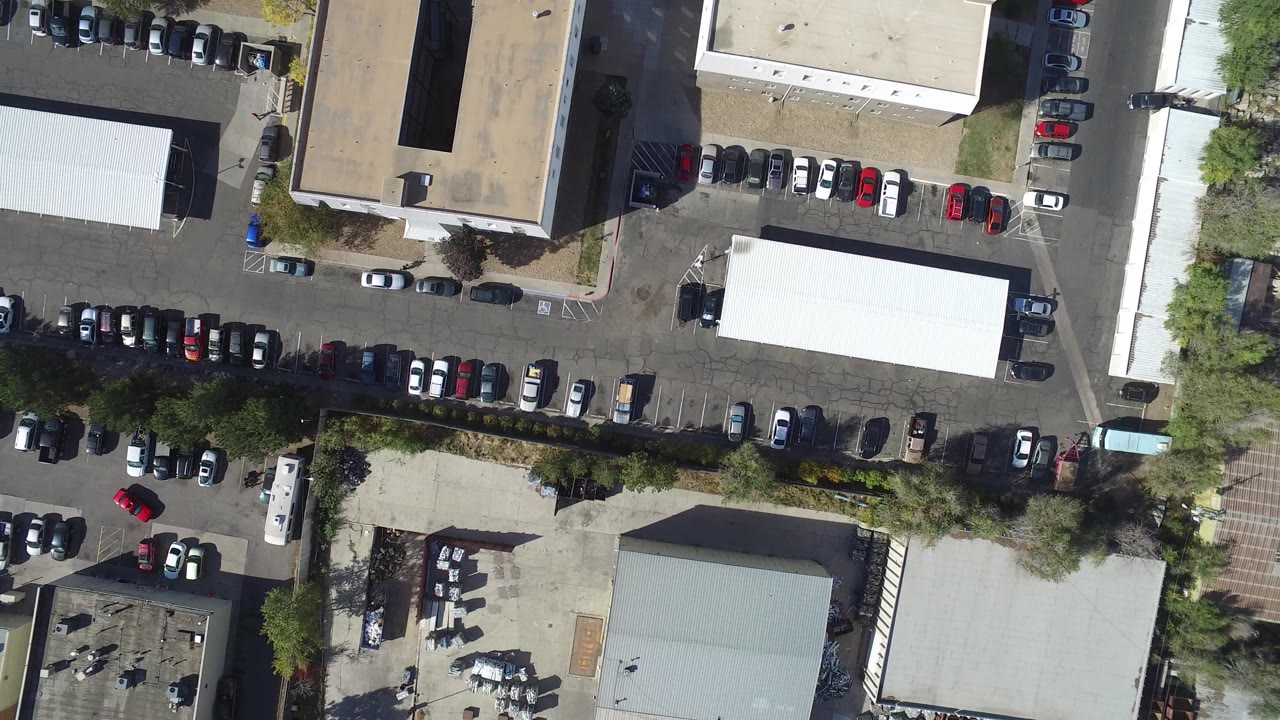 Overhead frame from Hidden Lake, Colorado showing parked cars around flat-roof commercial buildings, tree shadows, asphalt drive lanes, and no visible people.