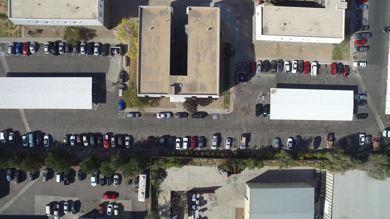 High overhead view in Hidden Lake, Colorado showing flat-roof buildings, rows of parked cars, access lanes, and no visible people at the opening moment.