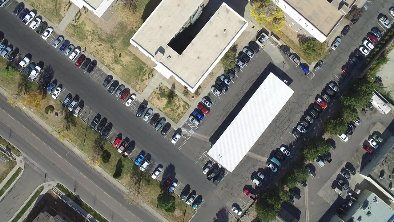 Opening overhead frame in Hidden Lake, Colorado, showing two flat-roofed buildings, packed parking rows, a curved road edge, trees, and no visible people.