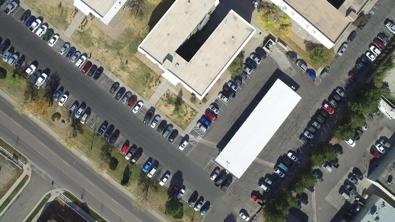 Aerial parking lot view with two light-roofed buildings and a road crossing the frame.