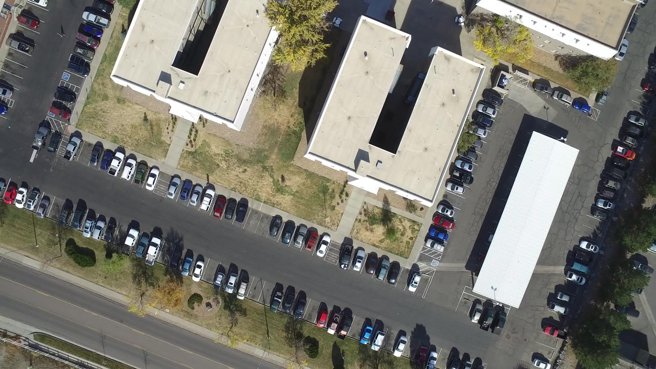 Overhead view of parked cars, flat-roofed buildings, road, grass, and trees.