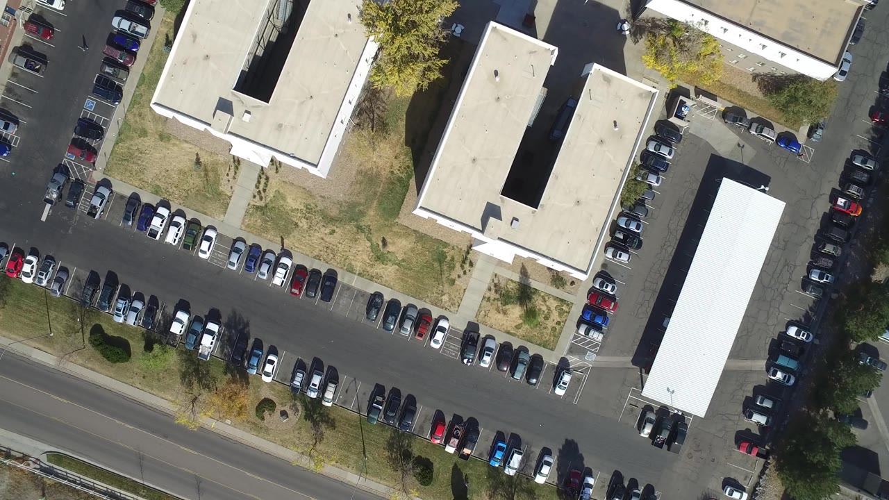 Vertical aerial frame in Hidden Lake, Colorado with a long line of parked cars, pale rooftops, grass strips, small trees, and no visible people.