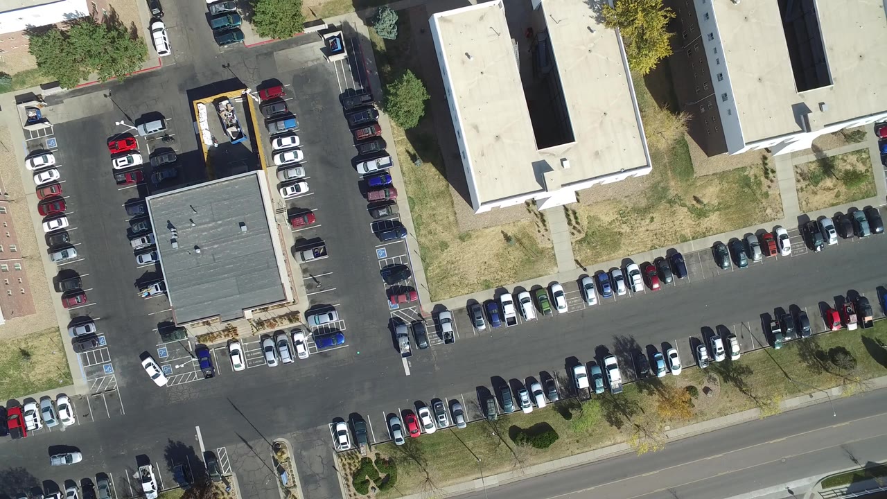 Overhead view of a parking lot with many cars and flat-roofed buildings.