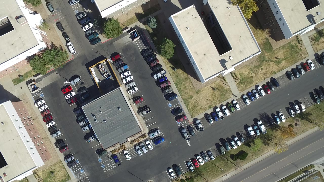 Overhead Hidden Lake, Colorado frame with a dark-roof building near the lower center, rows of parked cars, adjoining roads, sidewalks, dry grass, and no visible people.