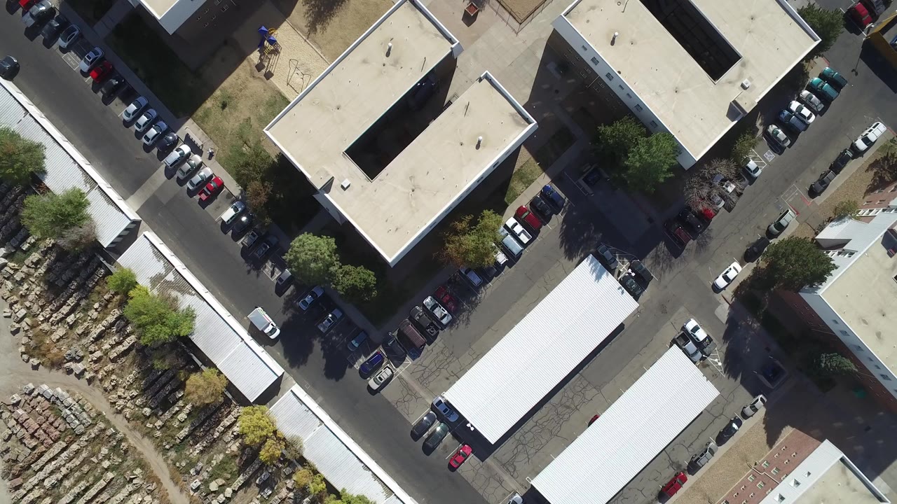 Top-down aerial view of streets, flat-roof buildings, parked cars, trees, and a materials yard.