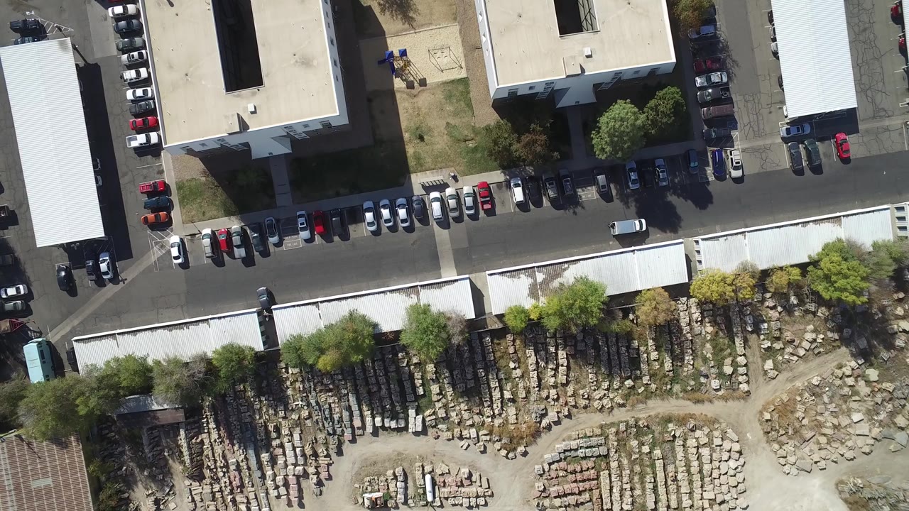Overhead view of parked cars, warehouse roofs, a road, and a material yard.