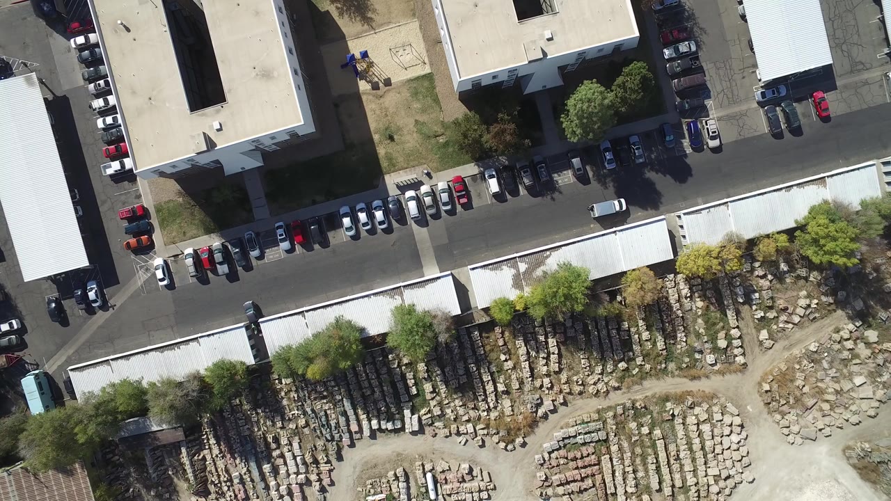 Near-vertical aerial frame over Hidden Lake, Colorado showing office building roofs, a row of parked cars, a paved road, trees, and stacked rock materials with no visible people.