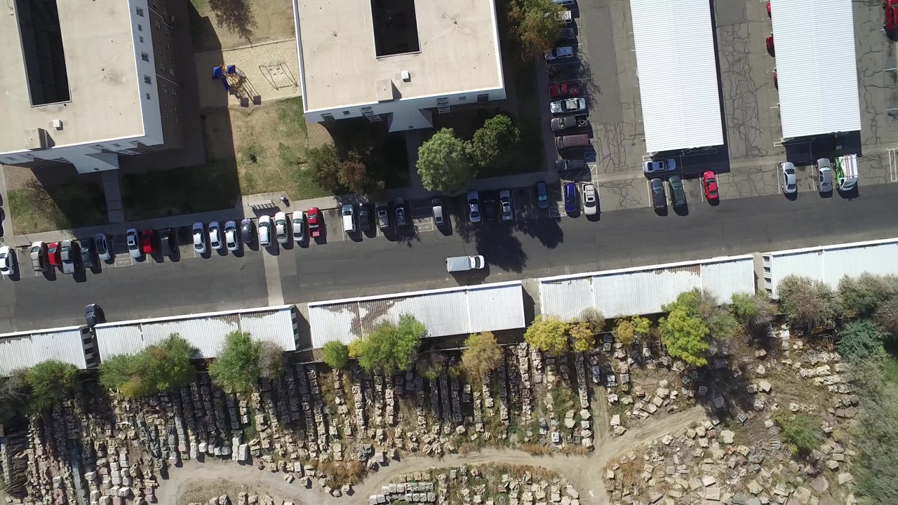 Top-down aerial view of a road, parked cars, buildings, trees, and rows of stone markers.