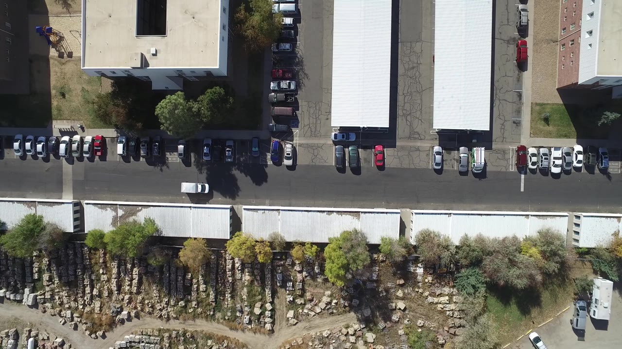 Top-down aerial view of a road, parked cars, rooftops, trees, and outdoor storage rows.