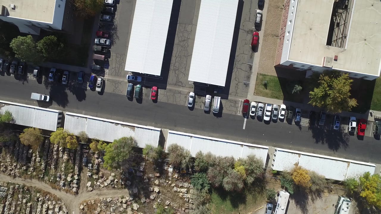 Aerial frame over Hidden Lake, Colorado showing a paved road between light-roofed buildings, parked cars, trees, and a rocky yard area, with no visible people.