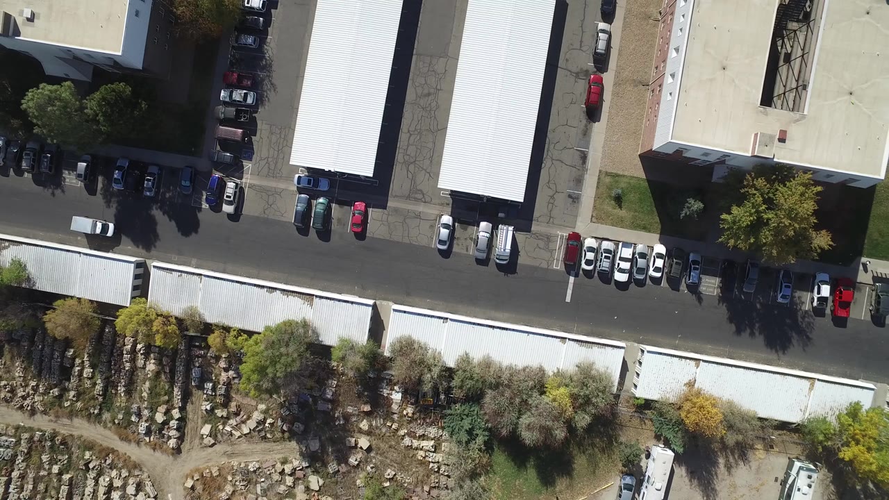 Aerial frame over Hidden Lake, Colorado with parked vehicles lining the road, white-roofed structures, tree shadows, and rough landscaped ground, with no visible people.