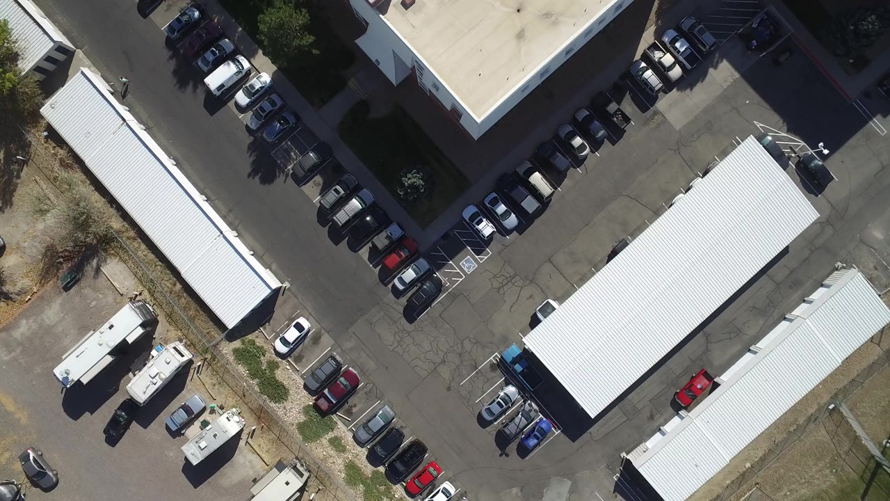 Overhead frame of a Hidden Lake, Colorado parking lot with white-roofed buildings, parked cars, trailers, dry ground, and no visible people.