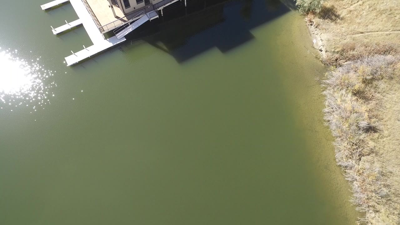 Overhead Hidden Lake, Colorado view with sun glare on the left, two small dock sections near the upper edge, a dark house shadow across the water, and no visible people.