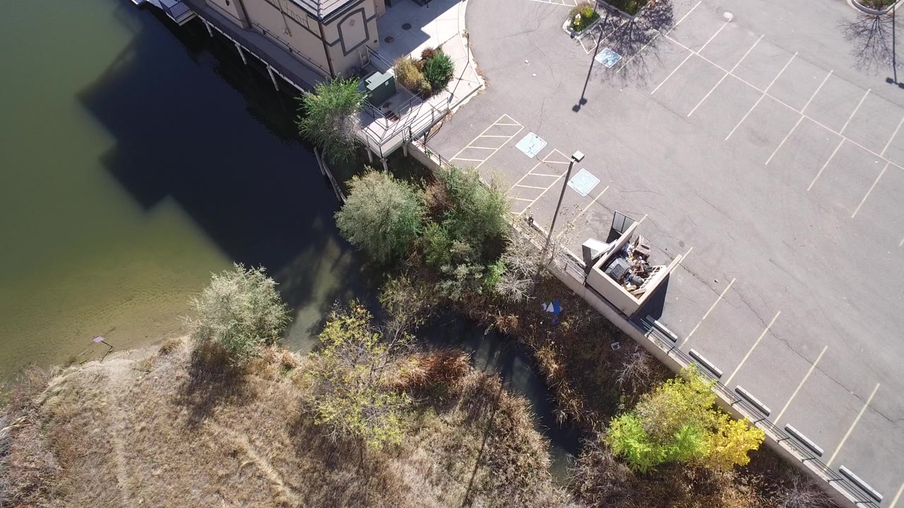 Aerial frame over Hidden Lake, Colorado showing green water, shoreline brush, an empty paved parking lot, accessible space markings, and no visible people.
