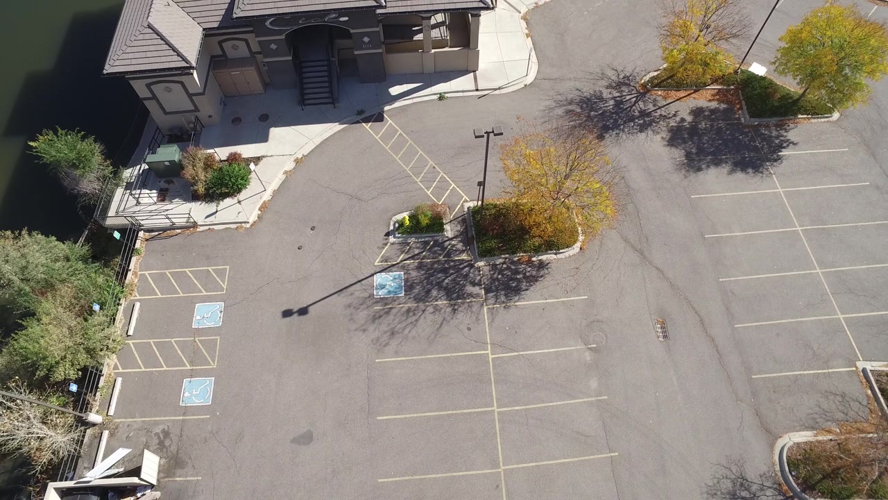Drone view of a paved lot with trees and a building along the top edge.