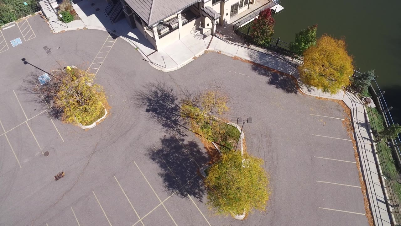 Aerial view over Hidden Lake Colorado with empty marked parking spaces, autumn trees in planted islands, a building corner at the top, and water running down the right side, with no visible people.