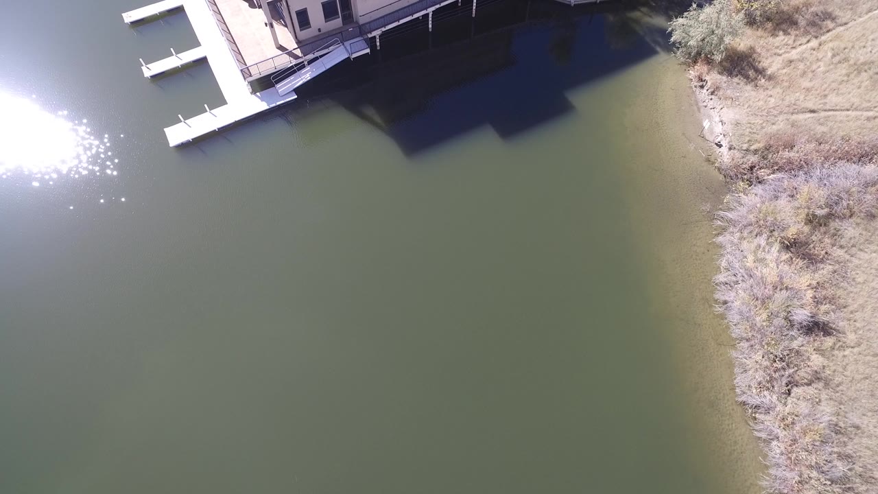 Aerial frame from Hidden Lake, Colorado with the house corner at the top, docks extending into green water, dry shoreline vegetation on the right, and no visible people.