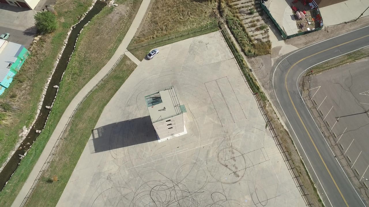 Aerial frame over Hidden Lake, Colorado with the small patio structure closer to center, tire or scuff marks on the concrete, a curved road at right, and a thin water channel along the grass, with no clearly visible people.