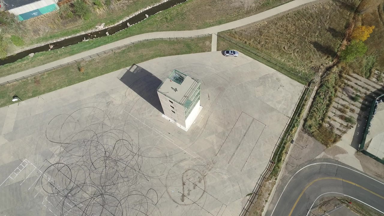 Top-down aerial view of a tower, marked concrete, paths, creek, and parked car.