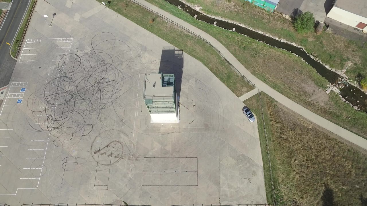 Overhead view of a concrete lot, small building, road, grass, and creek.